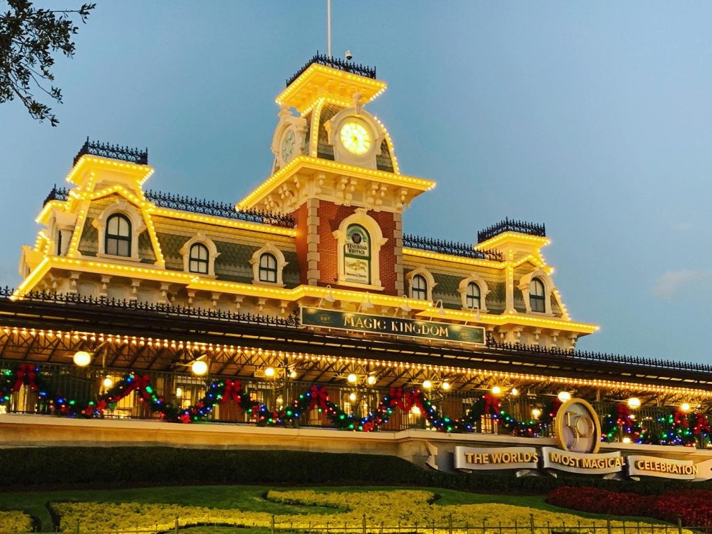 On the approach to Magic Kingdom during winter, the Main Street train station is lit up with festive, white, lights, with colorful lights on hanging wreaths across the front fence.