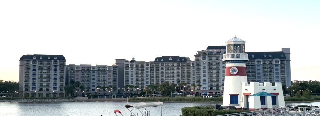 A full view of the Riviera resort at Disney World, taken from Caribbean Beach Resort. There's a white and red lighthouse in the foreground from Caribbean Beach, with the darker grays and blues of the Riviera resort in the background.